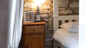 A bedroom with a stone wall, a wooden nightstand with a lamp, a framed picture, a bunk bed, and a white curtain.