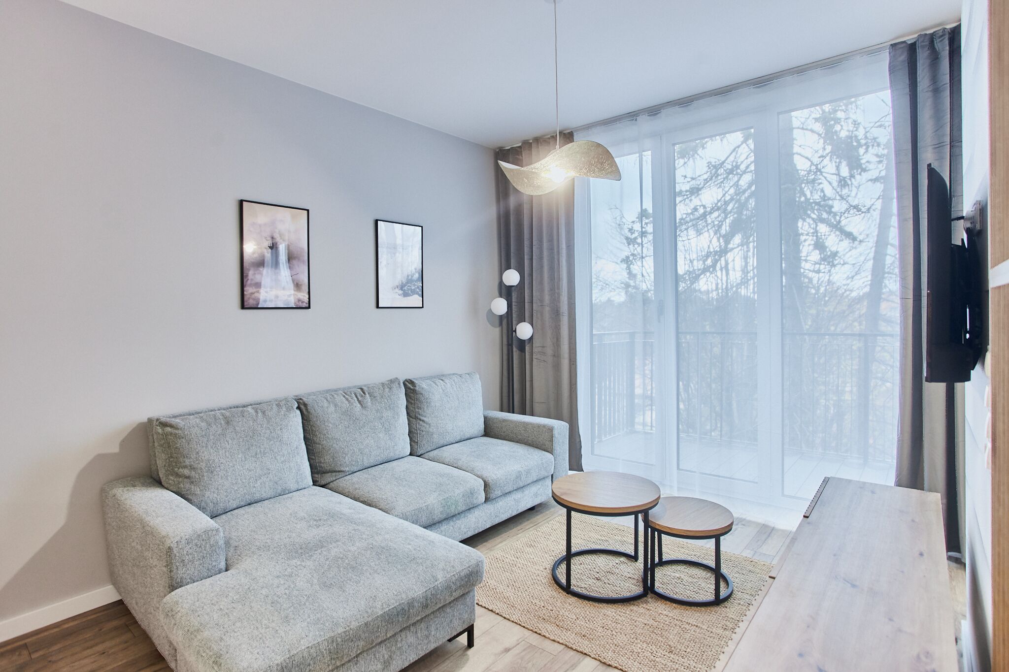 Ein modernes Wohnzimmer mit einer grauen Eckcouch, zwei kleinen runden Tischen, einem großen Fenster mit Vorhängen und zwei gerahmten Kunstwerken an der Wand.