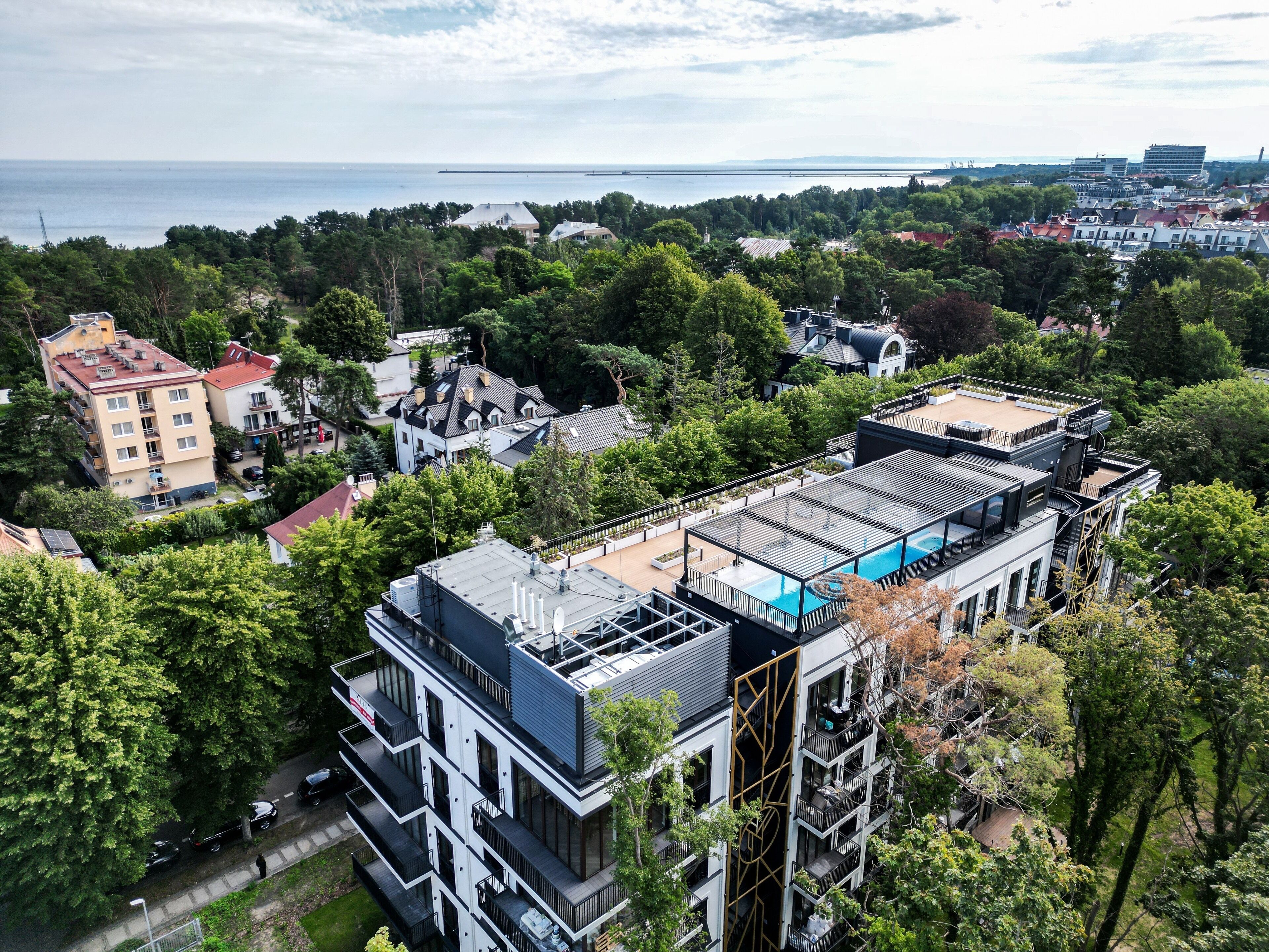 A modern multi-story building with a rooftop pool, surrounded by greenery and other residential buildings.