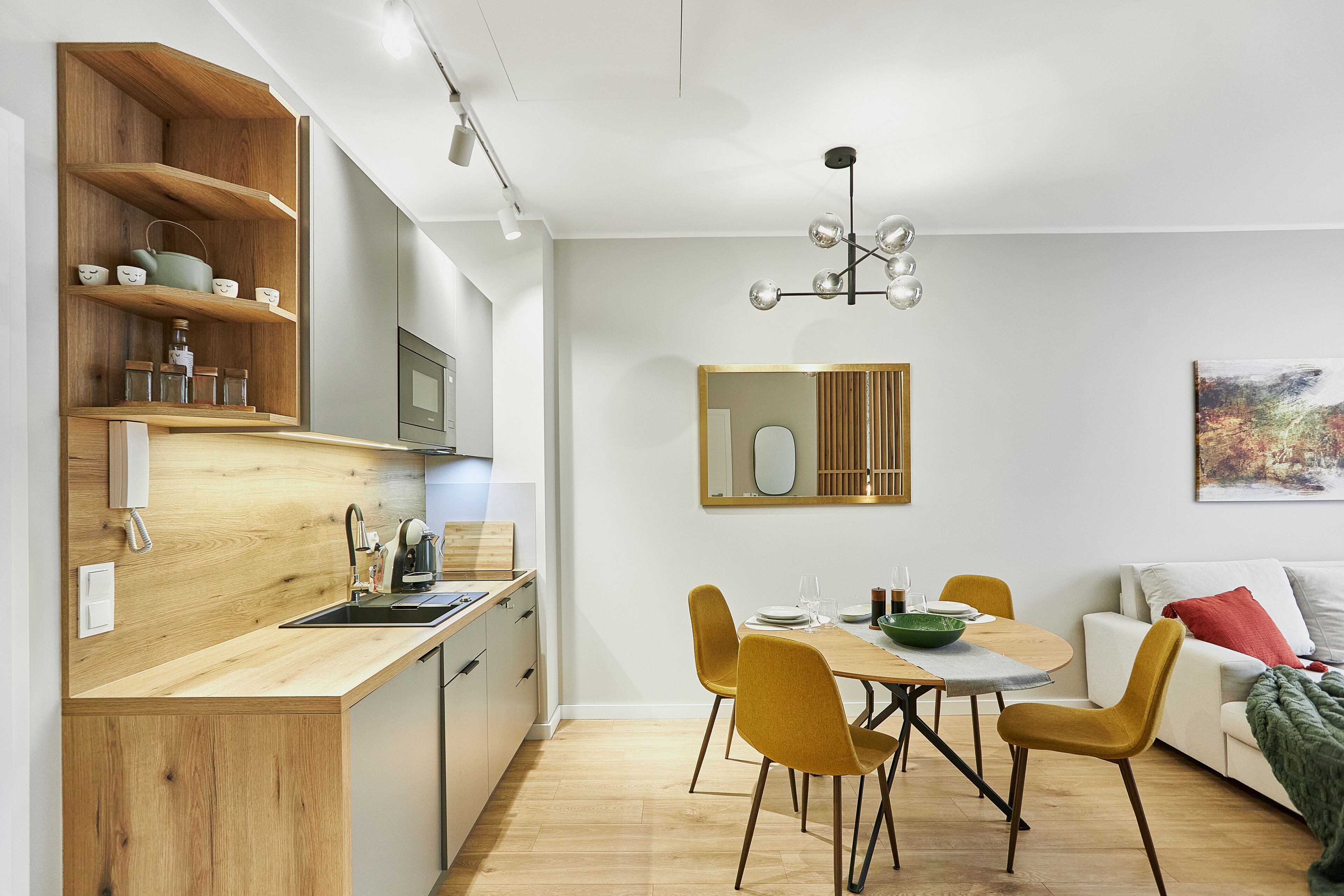 A modern kitchen with wooden shelves, a dining area with yellow chairs, and a white sofa with a red pillow.