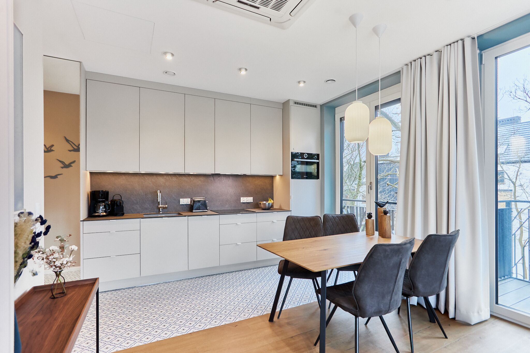 A modern kitchen with white cabinets, a wooden dining table, and a large window with curtains.