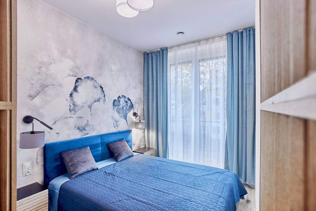 A modern bedroom with a large bed, a wall-mounted lamp, and a view of the outdoors through sheer curtains.