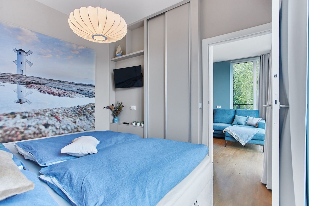 A bedroom with a bed, a TV, a painting of a lighthouse, and a view of a blue sofa in the adjacent room.