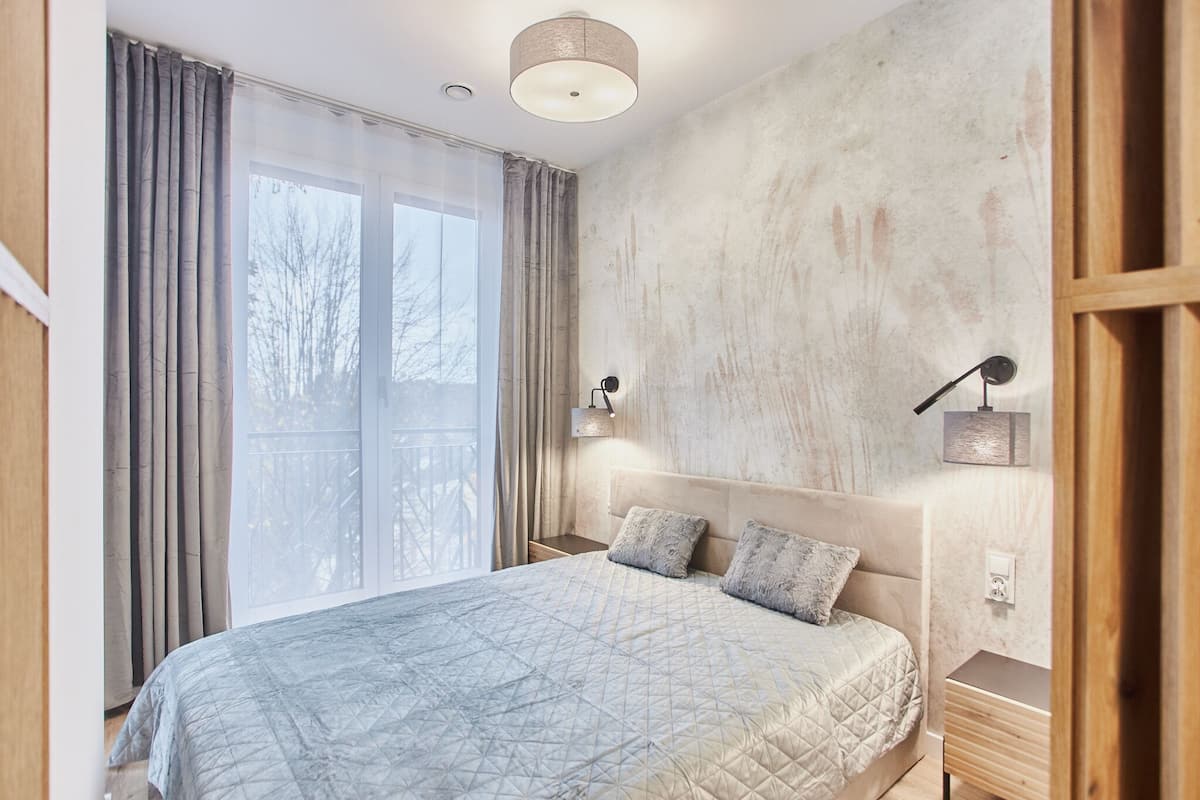 A bedroom with a large bed, two bedside lamps, a wooden nightstand, and a view of trees through the window.