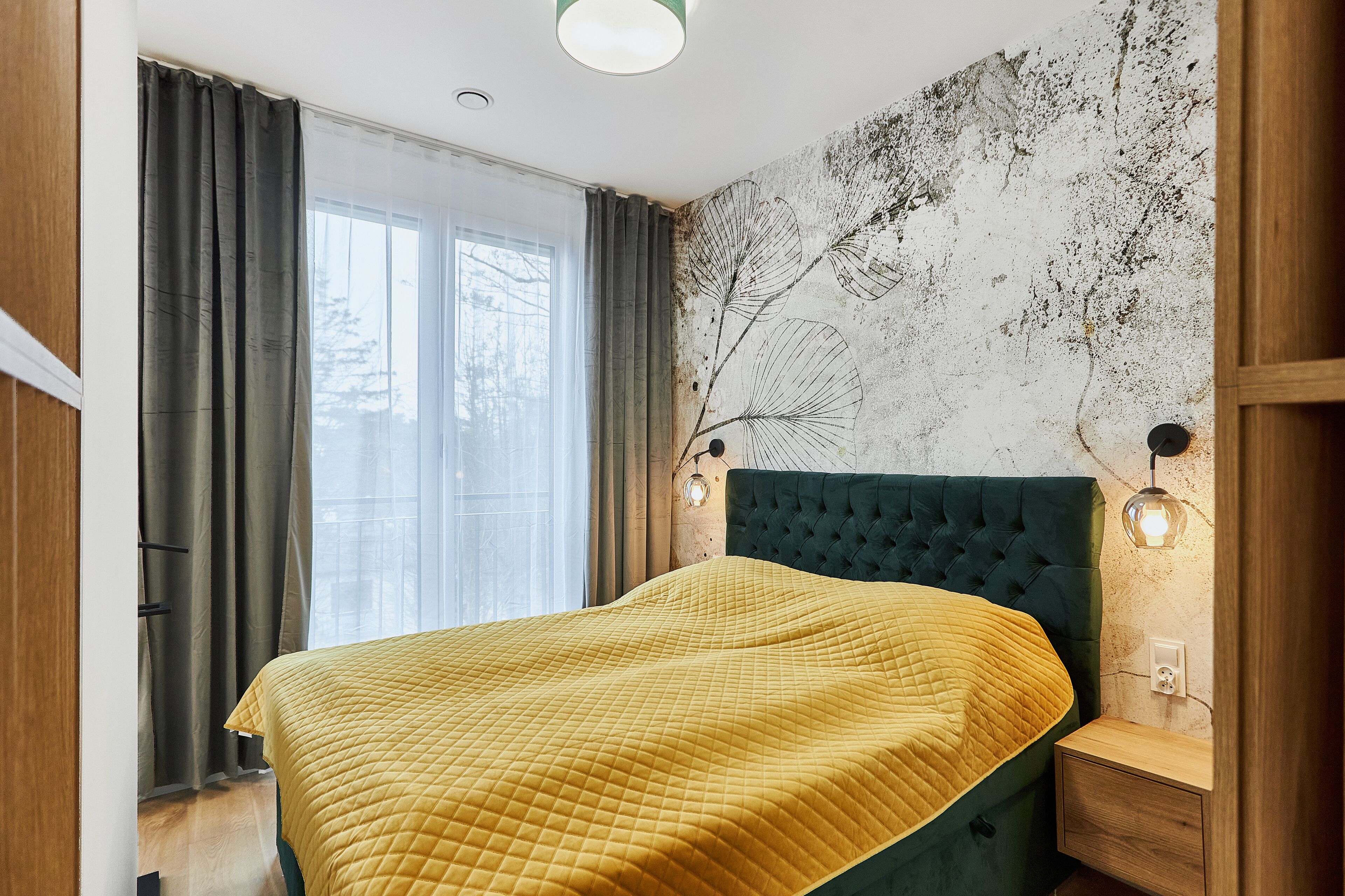 A bedroom with a large bed, a wooden nightstand, a wall-mounted lamp, and a window with curtains.