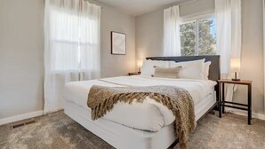A bedroom with a large bed, white curtains, a bedside table with a lamp, and a framed picture on the wall. - Mountain view | Games, Patio and BBQ in CO Springs (Colorado Springs)