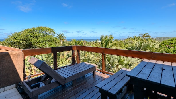 Une terrasse en bois avec deux bancs et une table, offrant une vue sur un paysage tropical avec des palmiers et sur l’océan.