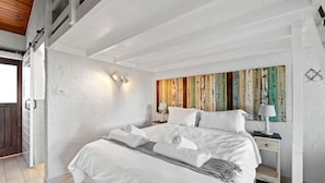 Une chambre spacieuse avec un grand lit, des murs blancs et une tête de lit en bois. On y trouve une table de chevet avec une lampe et un tableau accroché au mur.