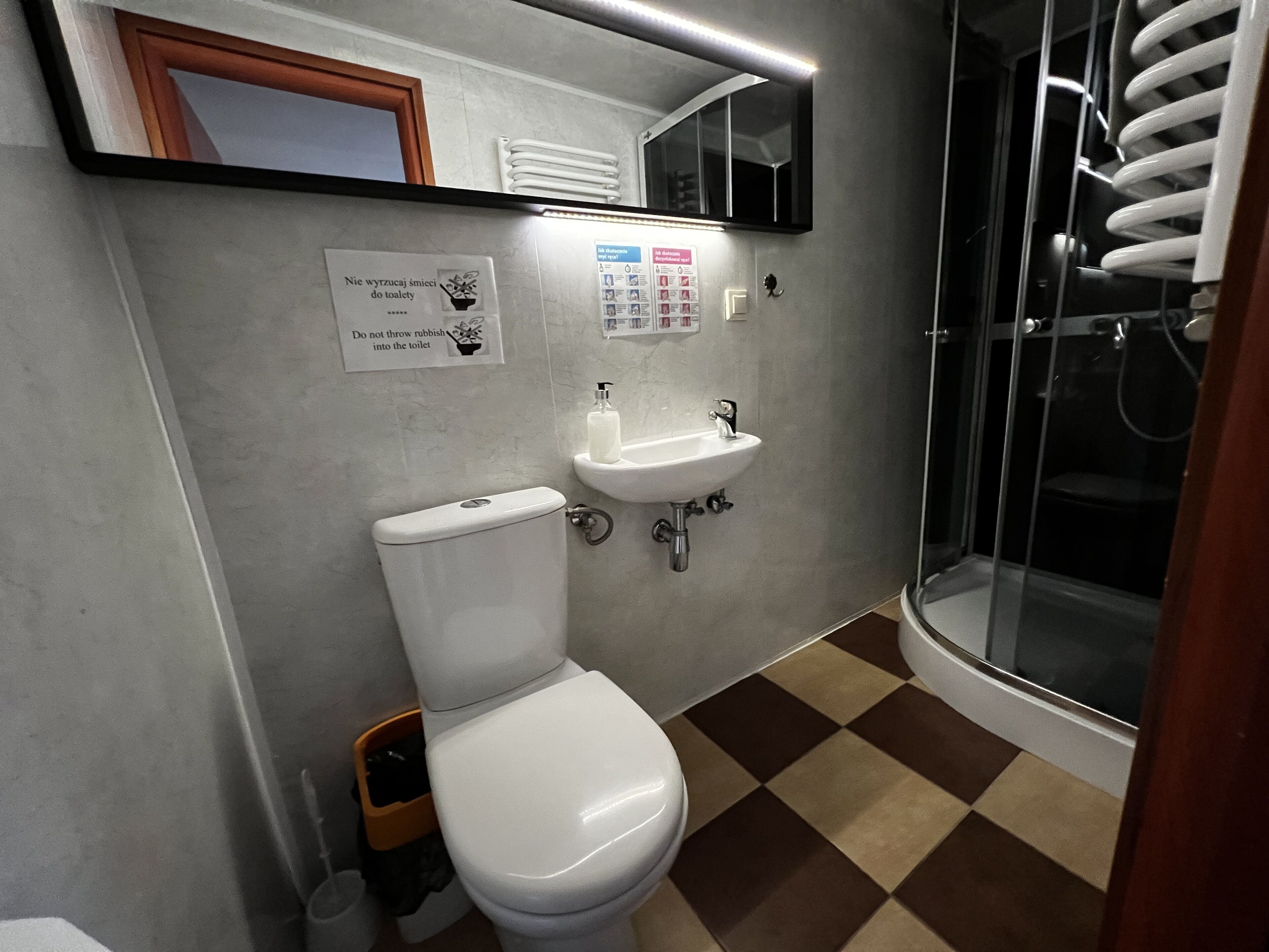 Un baño con inodoro, lavamanos y ducha. Hay un cartel en la pared y un cubo de basura.