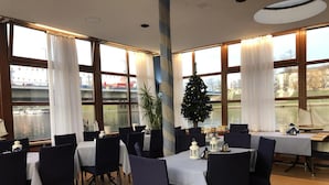 A dining area with tables set for four, large windows with curtains, a Christmas tree, and a view of a body of water. - The Boat - Hostel&Chill (Kraków)