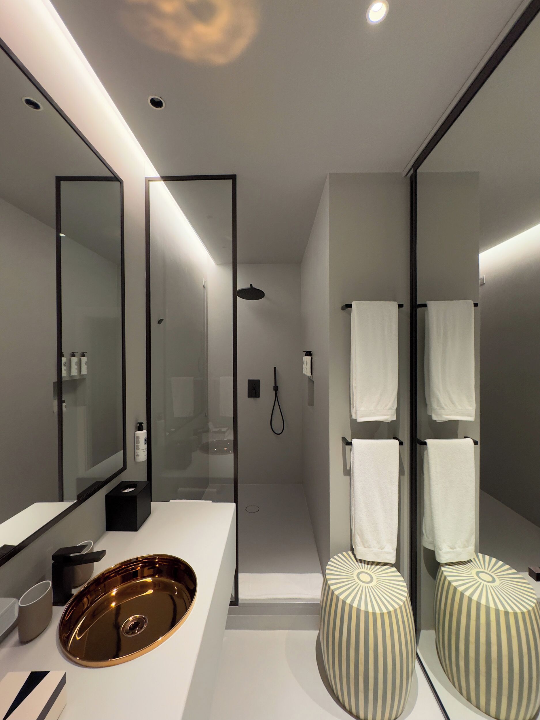 A modern bathroom with a large mirror, a glass-enclosed shower, and two striped ottomans.