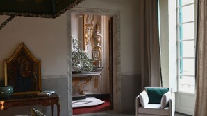 A hotel room with a large bed, a chair, a small table with a painting, and a view of a staircase and a door. - Palazzo Durazzo Suites (Genoa)