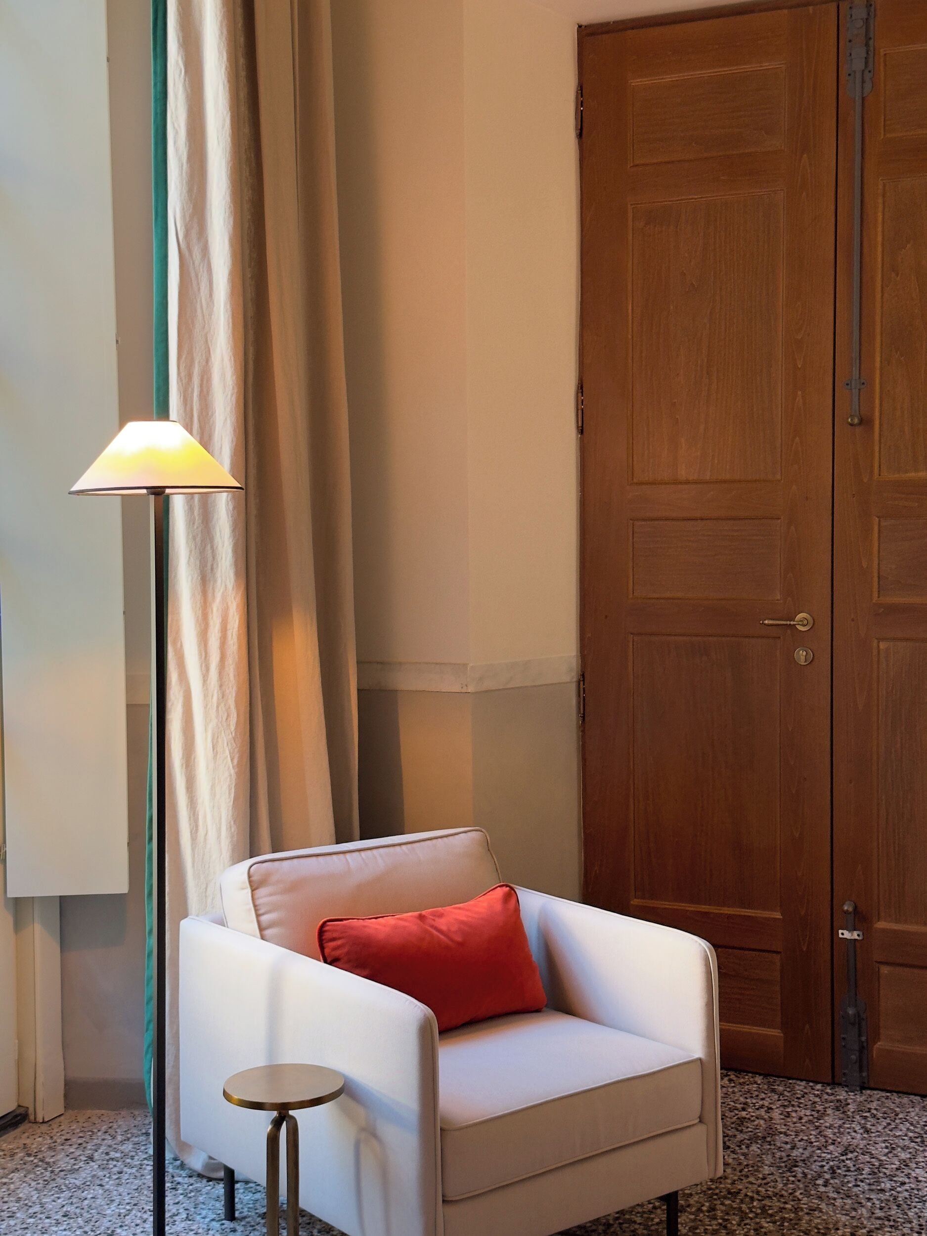 Een kamer met een witte fauteuil, een klein rond tafeltje, een vloerlamp en een houten deur.
