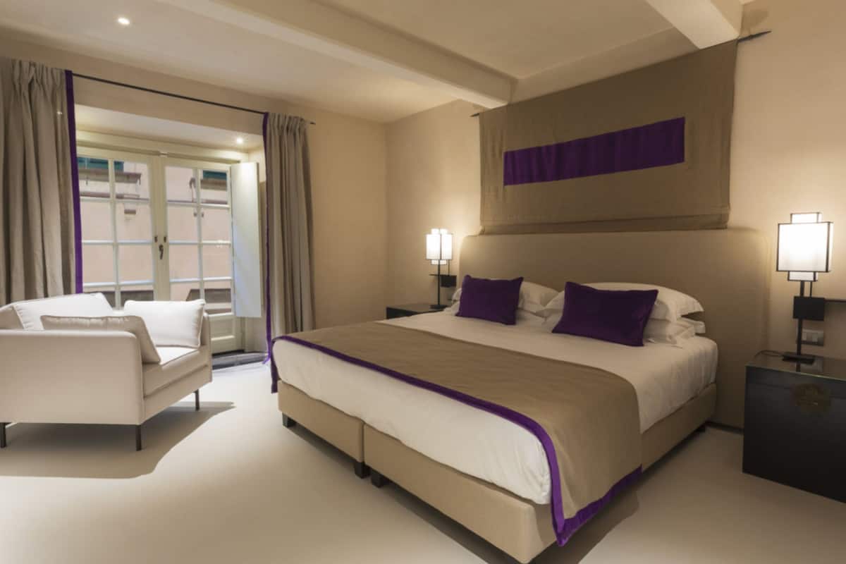 A modern hotel room with a large bed, two bedside tables with lamps, a sofa, and a view of the outside through a large window.