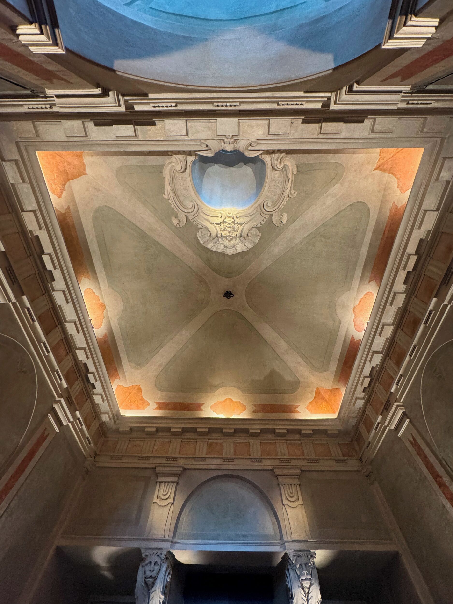 Een statige zaal met een versierd plafond en zuilen, een grote gewelfde doorgang, en een blauw dakraam.