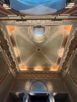 Een statige zaal met een versierd plafond en zuilen, een grote gewelfde doorgang, en een blauw dakraam.