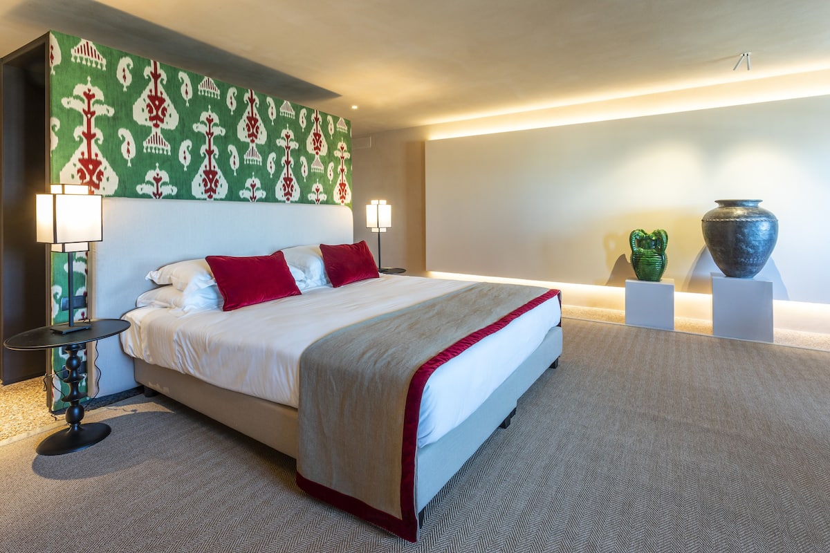 A hotel room with a large bed, two bedside lamps, a decorative headboard, and two vases on pedestals.