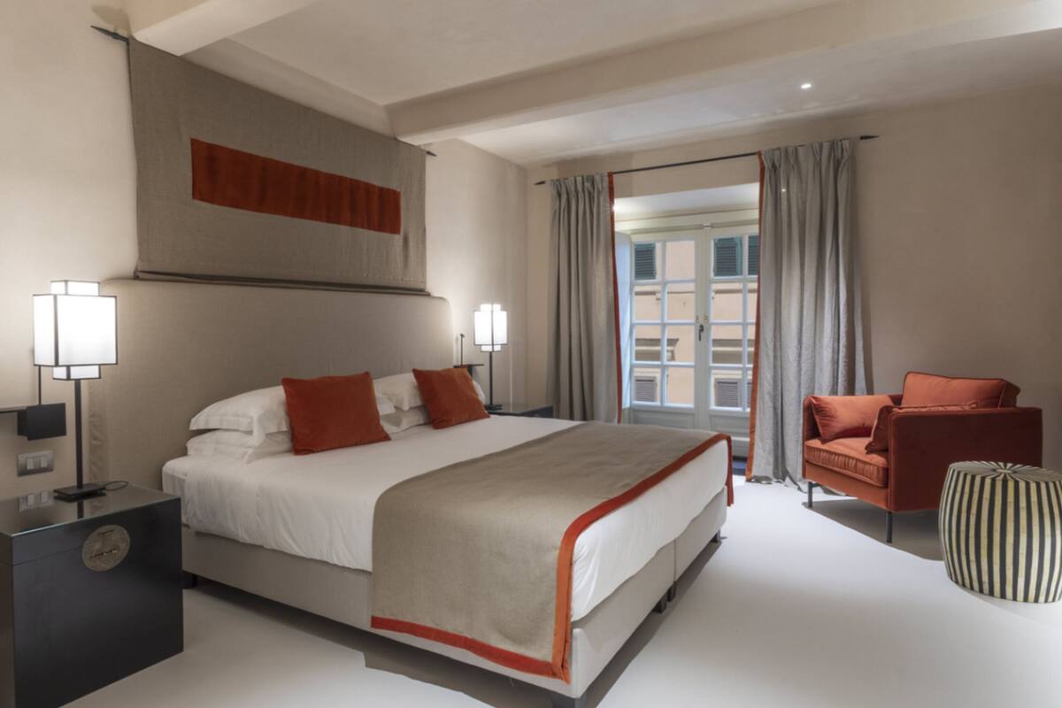 A modern hotel room with a large bed, two bedside lamps, a sofa, and a striped ottoman.