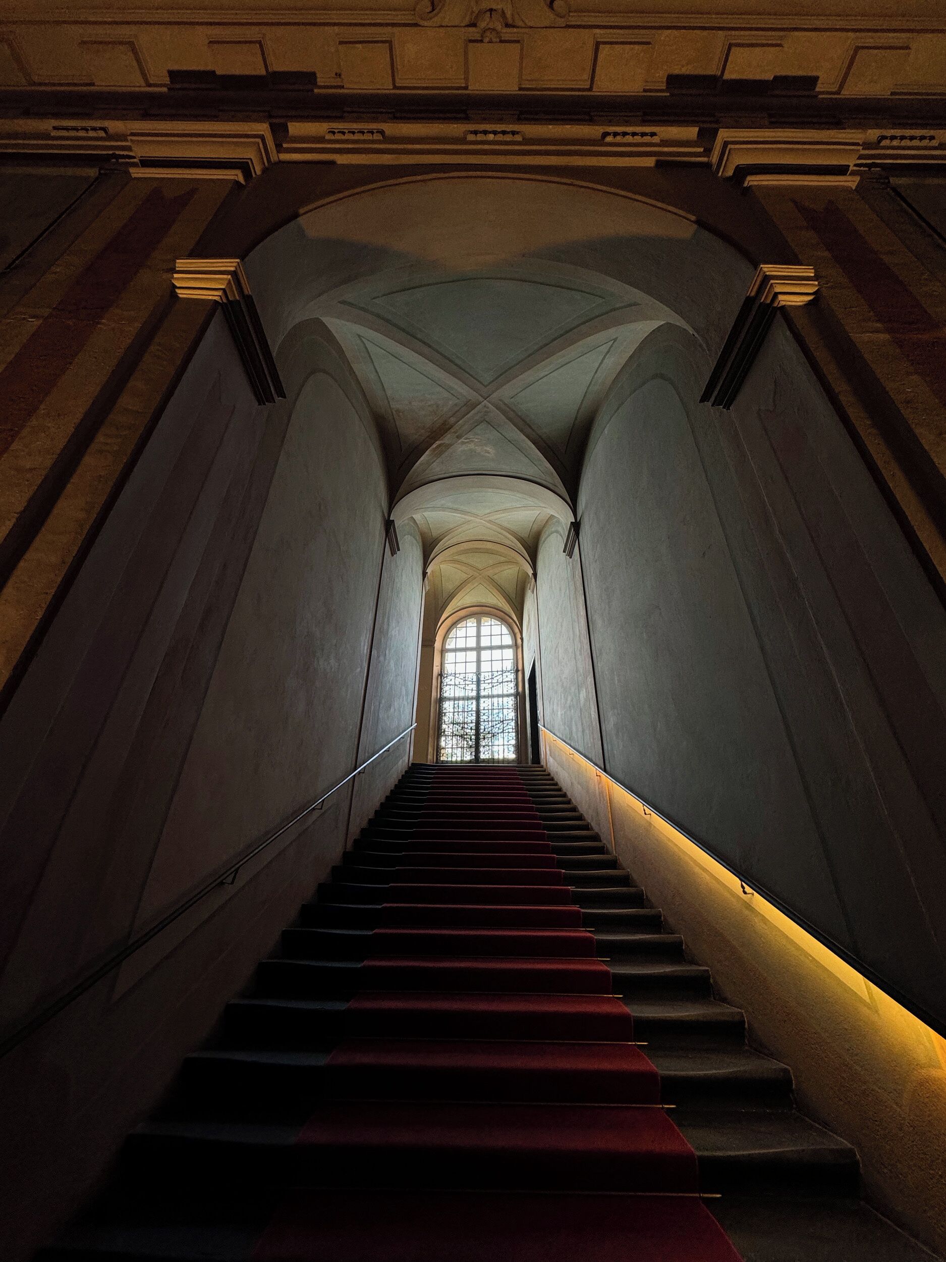 A grand staircase with a red carpet leading to a large arched window at the top.