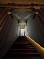 A grand staircase with a red carpet leading to a large arched window at the top.