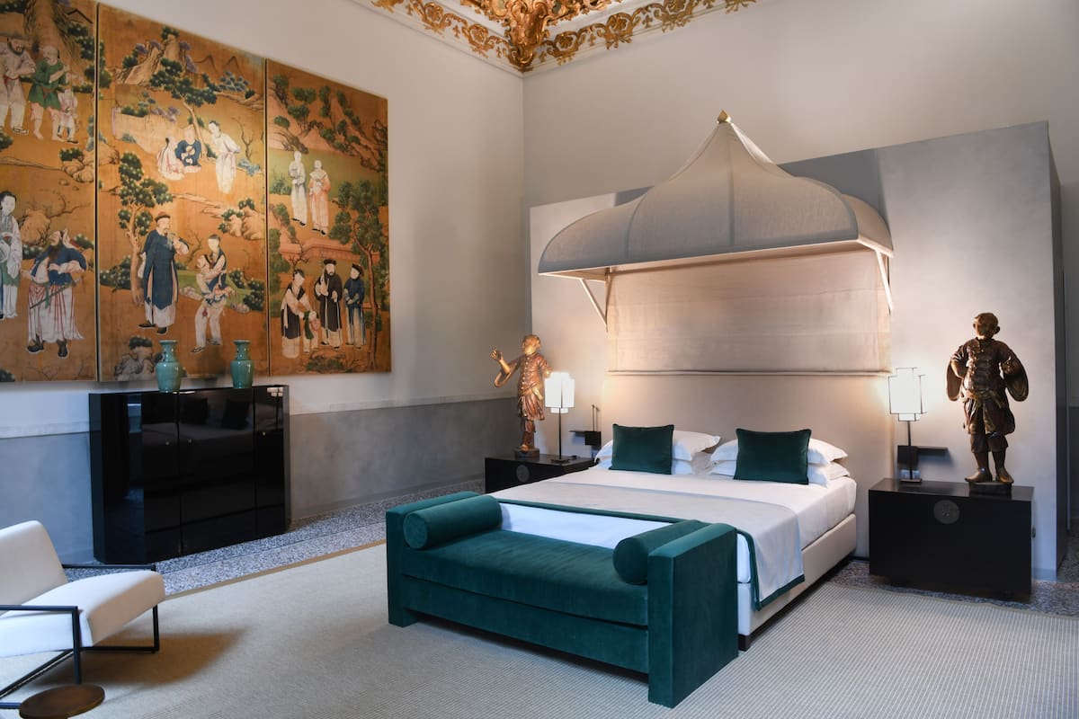 A modern hotel room with a large traditional painting, a canopy bed, a desk with a lamp, and a statue.