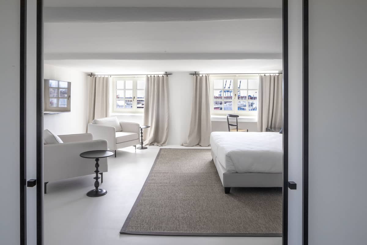 A hotel room with a bed, a sofa, a small table, and a view of the city through large windows.