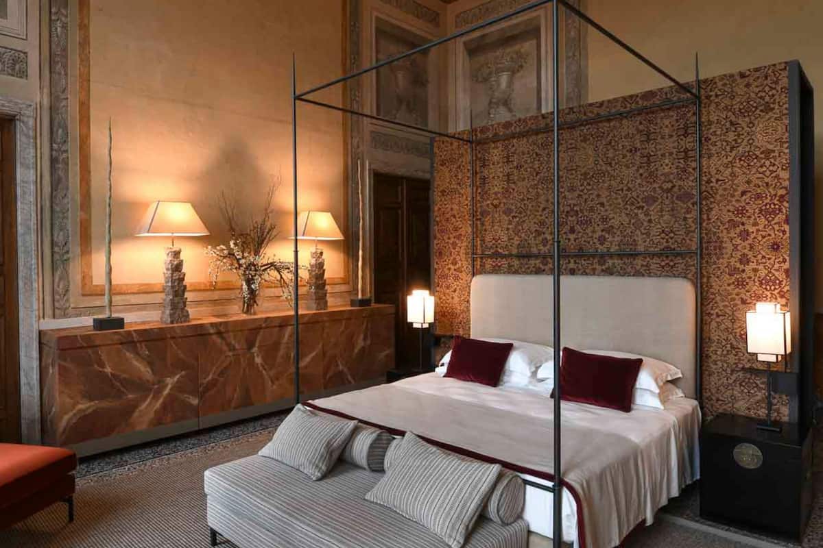 A four-poster bed with a marble headboard, a red upholstered bench, and a patterned wall.