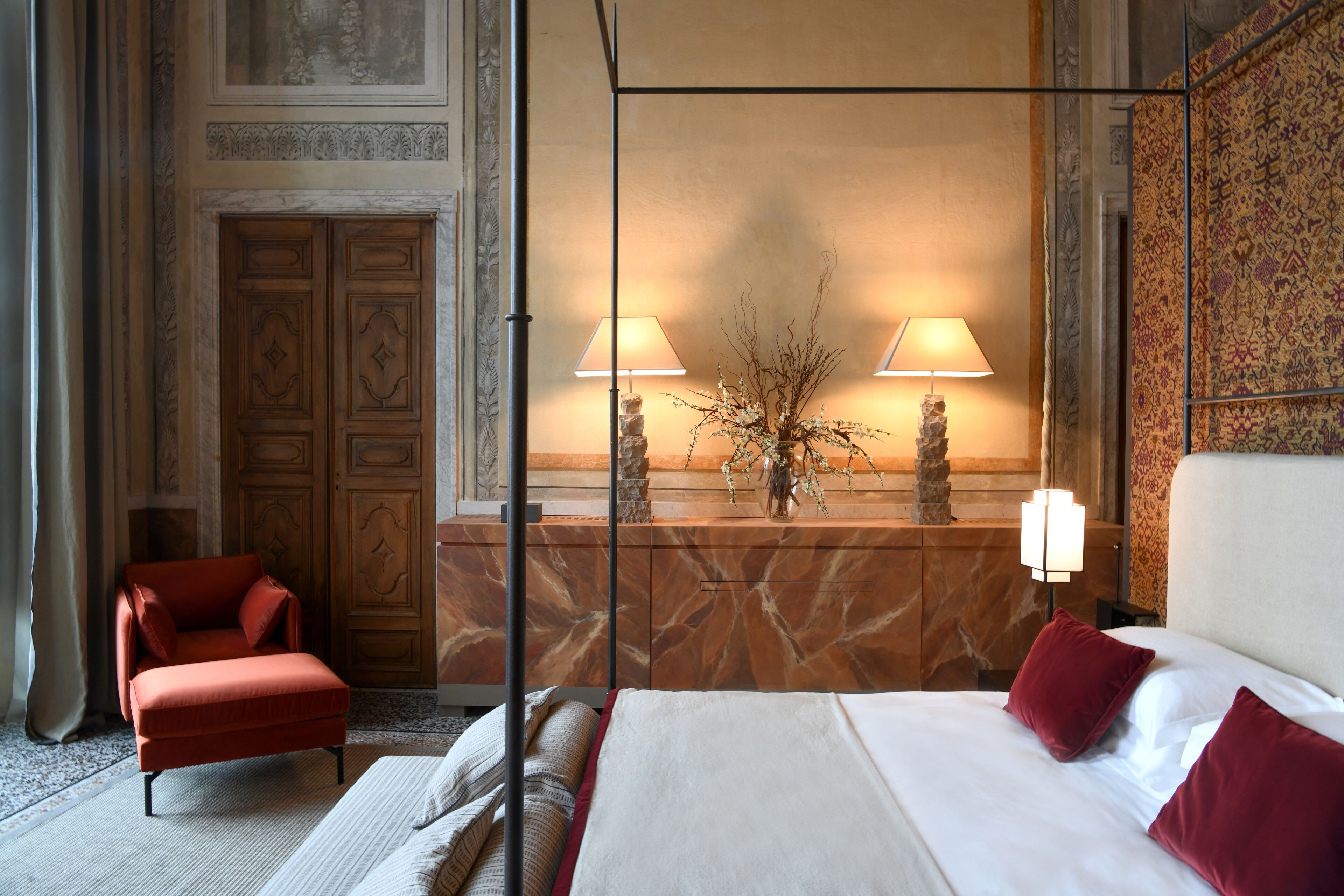 A hotel room with a bed, a chair, a nightstand with lamps, and a decorative wall.