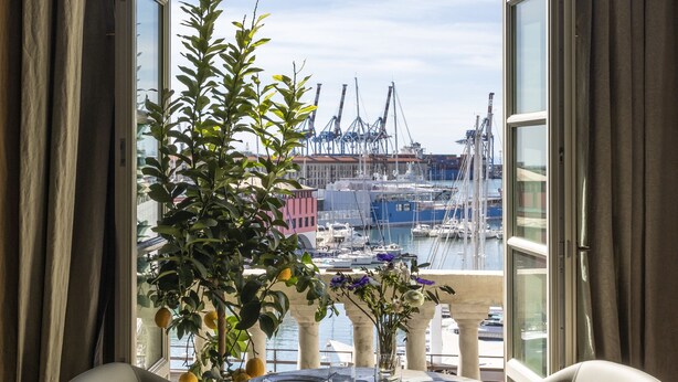 A dining area with a table and two chairs, a potted plant, and a view of a marina with boats and cranes. - Palazzo Durazzo Suites (Genoa)