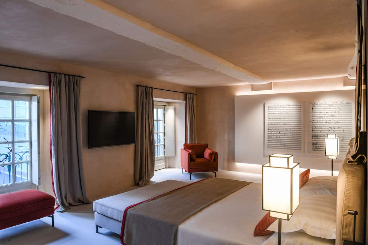 A hotel room with a large bed, a red armchair, a TV mounted on the wall, and a window with curtains.