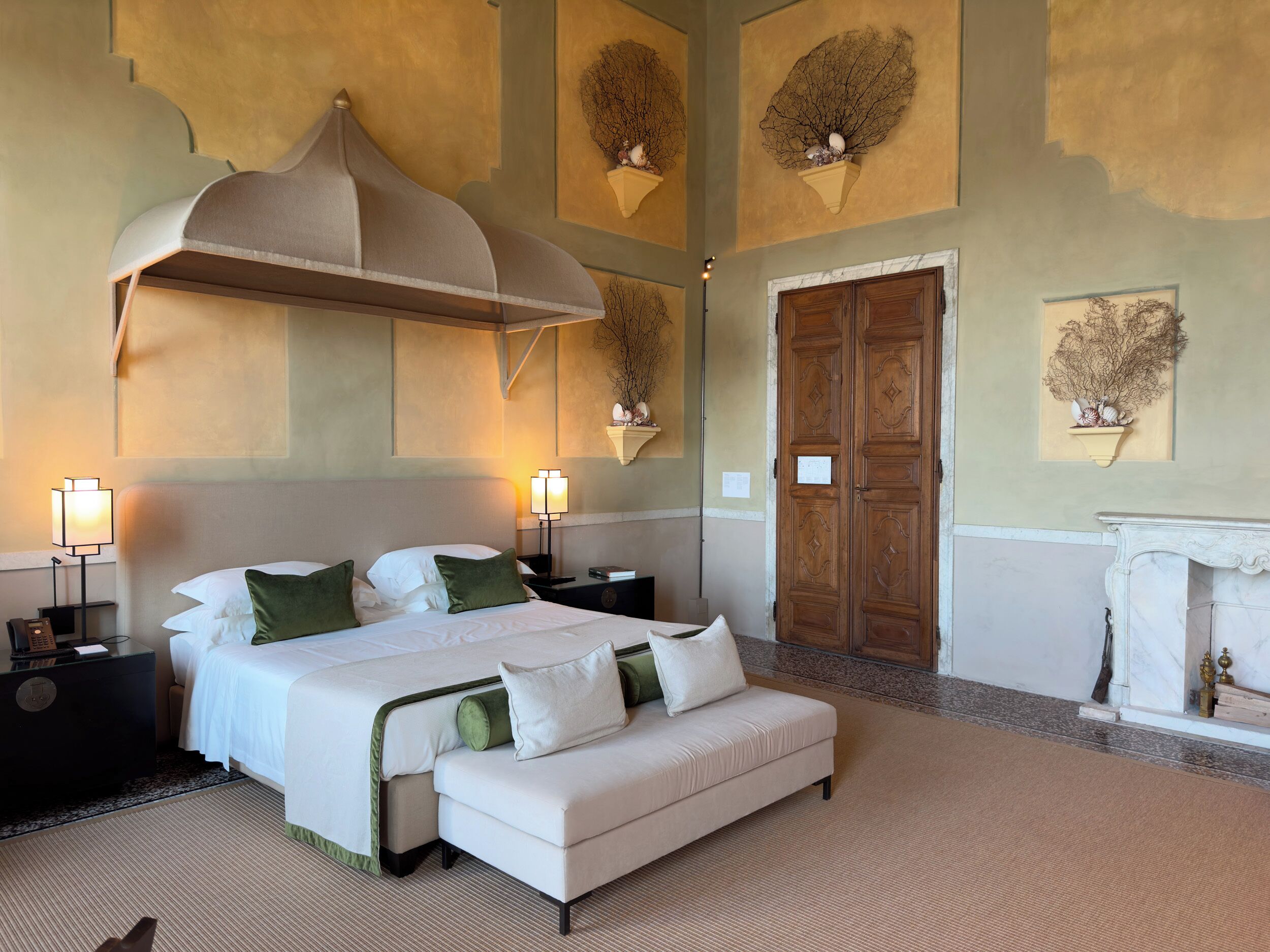 A hotel room with a large bed, two bedside tables, a nightstand, a wooden wardrobe, a fireplace, and wall-mounted sconces.