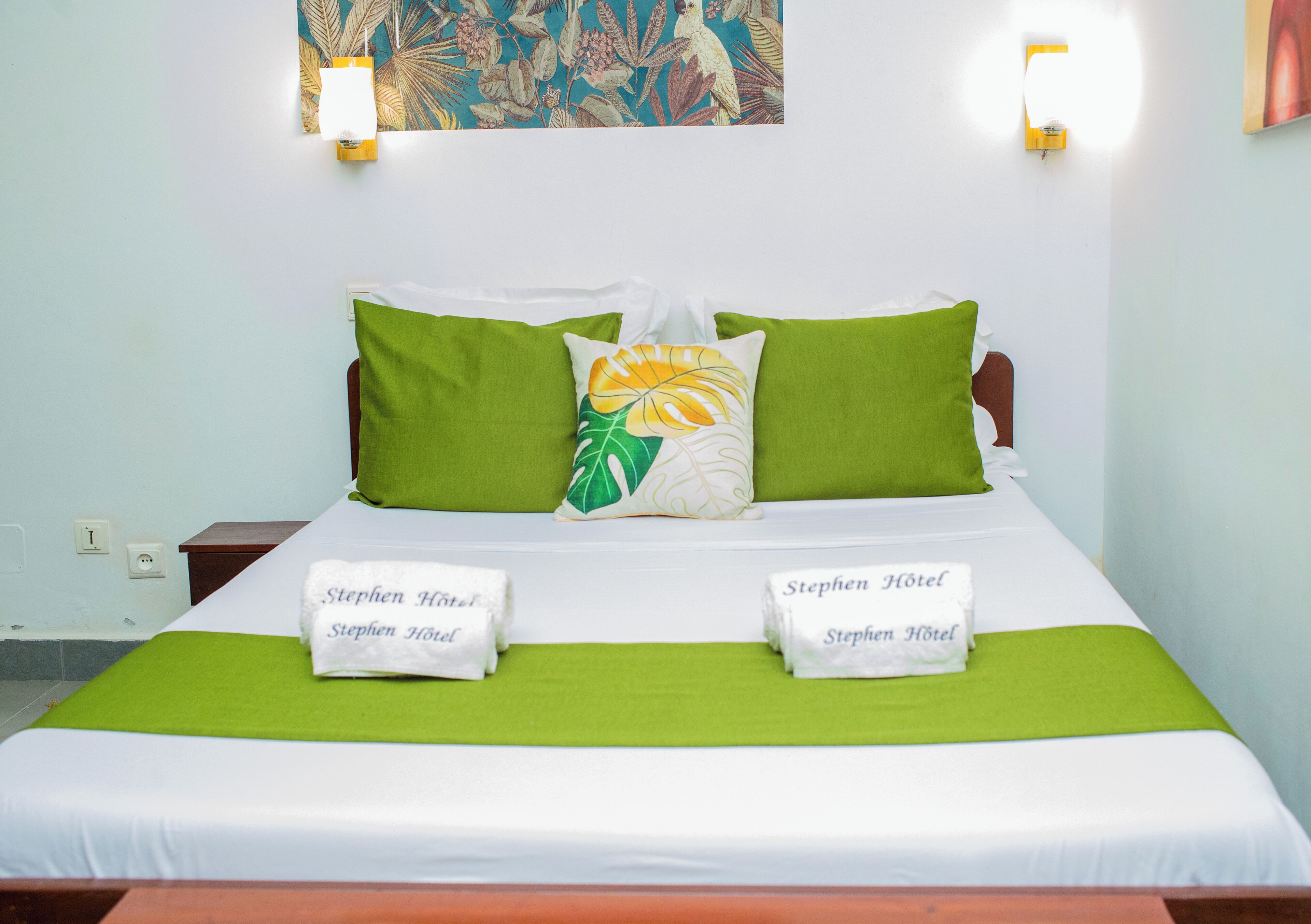 Cama arrumada com roupa de cama verde e branca, dois travesseiros e duas toalhas com a inscrição 'Stephen Hotel'.