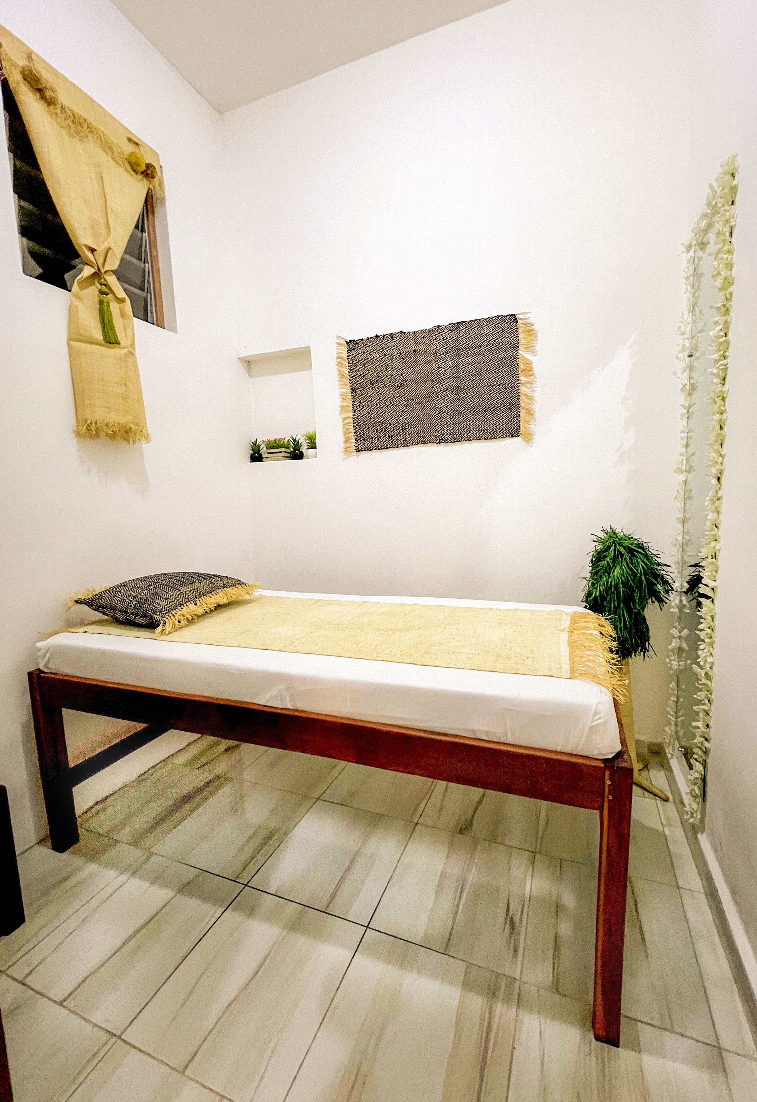 A small, clean room with a bed, a window with a curtain, a small shelf with plants, and a wall-mounted mirror.