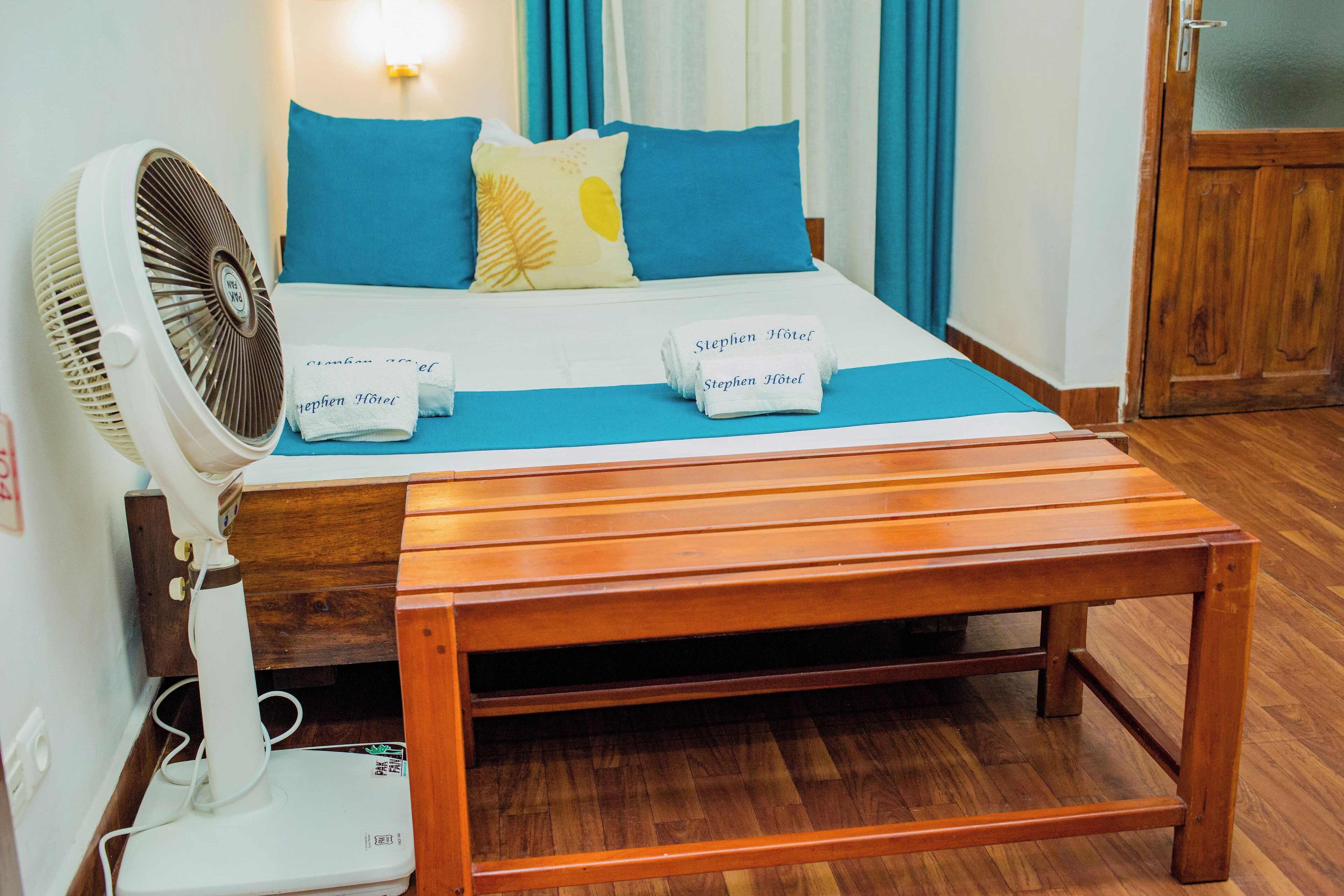 A neatly made bed with blue and yellow pillows, a white fan, and a wooden bench.