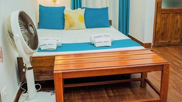 A neatly made bed with blue and yellow pillows, a white fan, and a wooden bench.