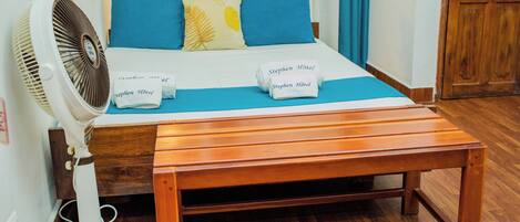 A neatly made bed with blue and yellow pillows, a white fan, and a wooden bench.