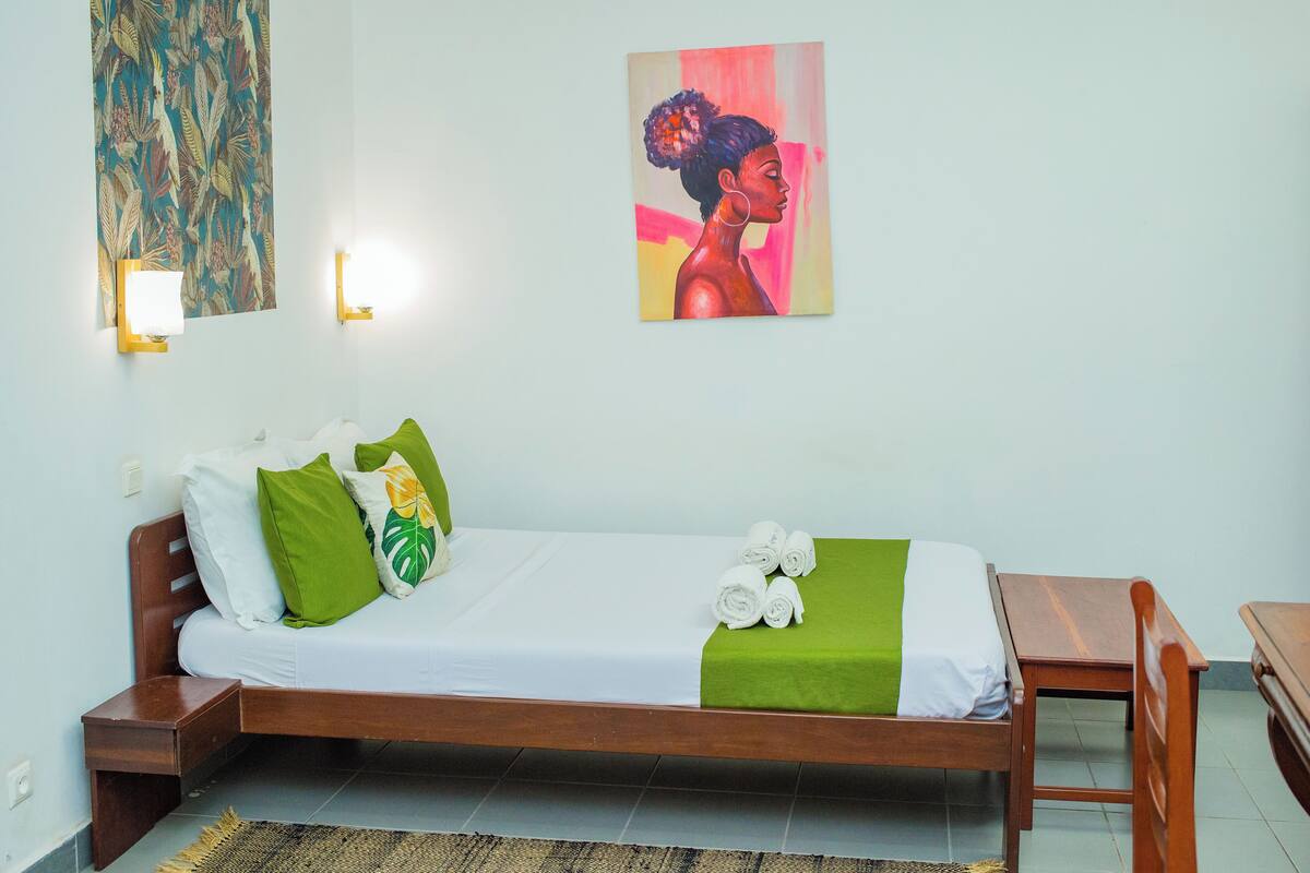 Ein Schlafzimmer mit einem hölzernen Bett, weißer Bettwäsche, grünen Kissen und einer grünen Tagesdecke. An der Wand hängt ein Gemälde und neben dem Bett steht ein kleiner Tisch.