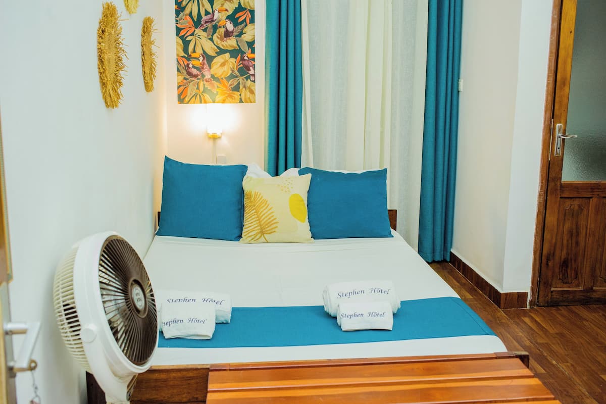 Ein ordentlich bezogenes Bett mit blauen und gelben Kissen, ein Ventilator und Handtücher mit dem Aufdruck "Stephen Hotel".