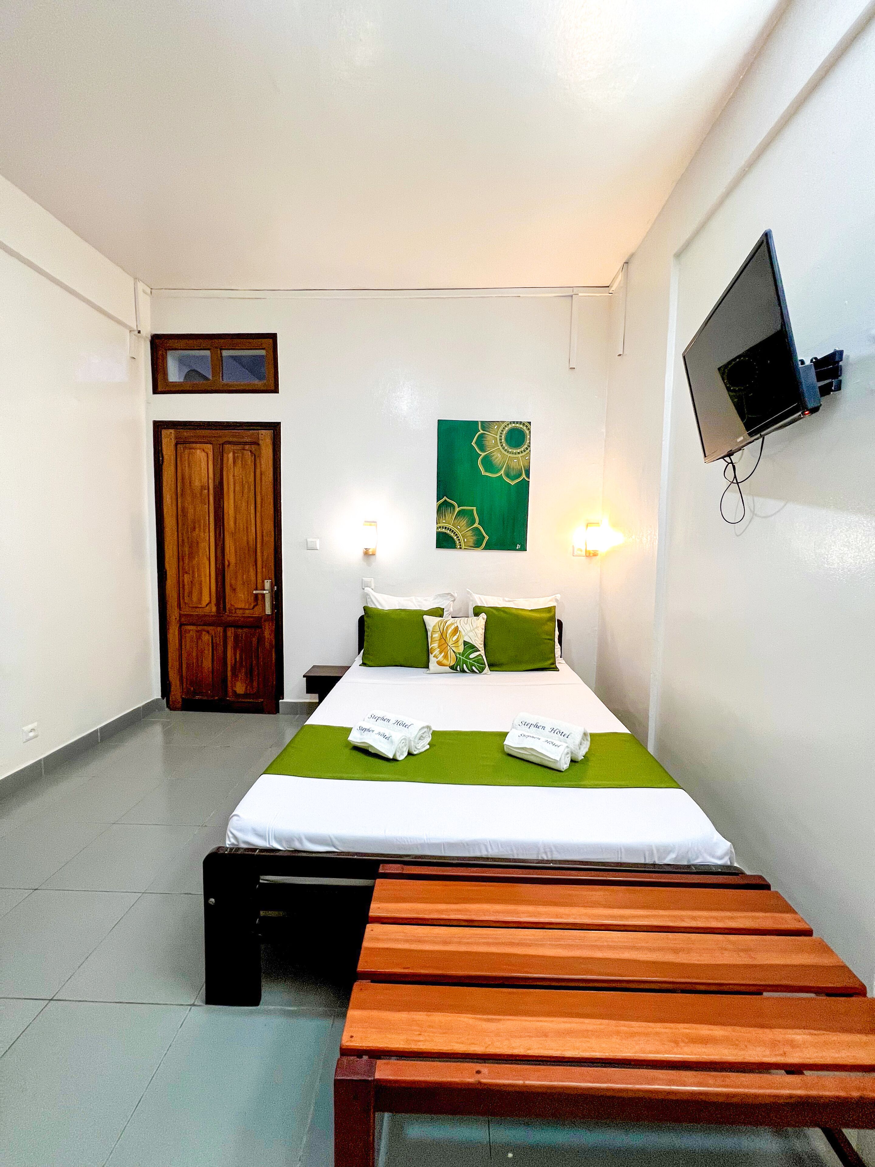 A hotel room with a bed, green and white bedding, a wooden bench, a TV mounted on the wall, and a door with a window.