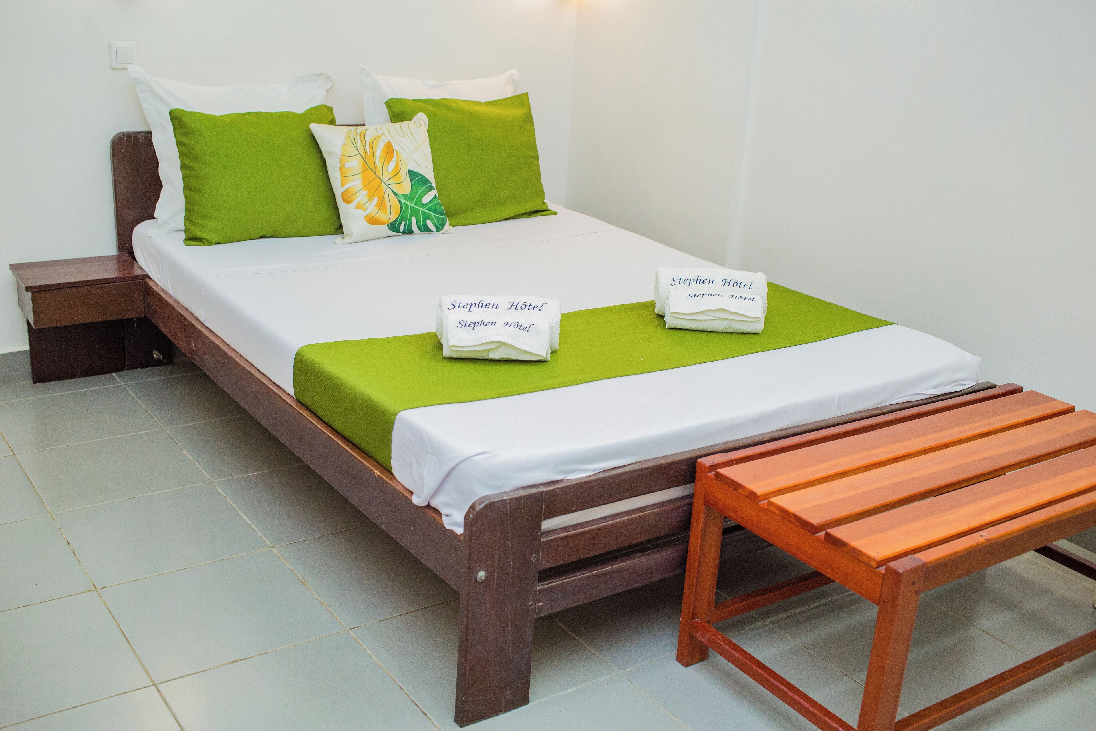 A neatly made bed with green and white bedding, two towels with text, a wooden bench, and a tiled floor.