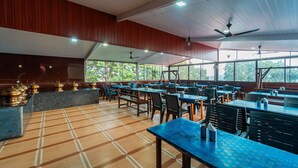 A spacious indoor dining area with multiple tables and chairs, large windows, and ceiling fans. - Itsy Hotels Sun N Shade (Karjat)