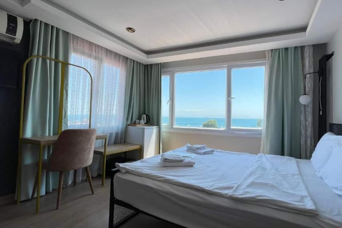 A hotel room with a bed, a desk, and a chair. There is a window with a view of the sea.
