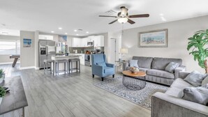Una sala de estar moderna con un sofá gris, un sillón azul y una mesa de centro. Hay una zona de cocina con armarios blancos y una mesa de comedor.
