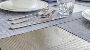 Una mesa de comedor preparada con platos, tazones, cubiertos y vasos, ubicada cerca de una ventana con vistas a árboles.
