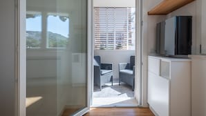 Une chambre moderne dotée d’une porte-fenêtre coulissante donnant sur un balcon où se trouvent deux fauteuils en osier et un téléviseur posé sur une étagère en bois.