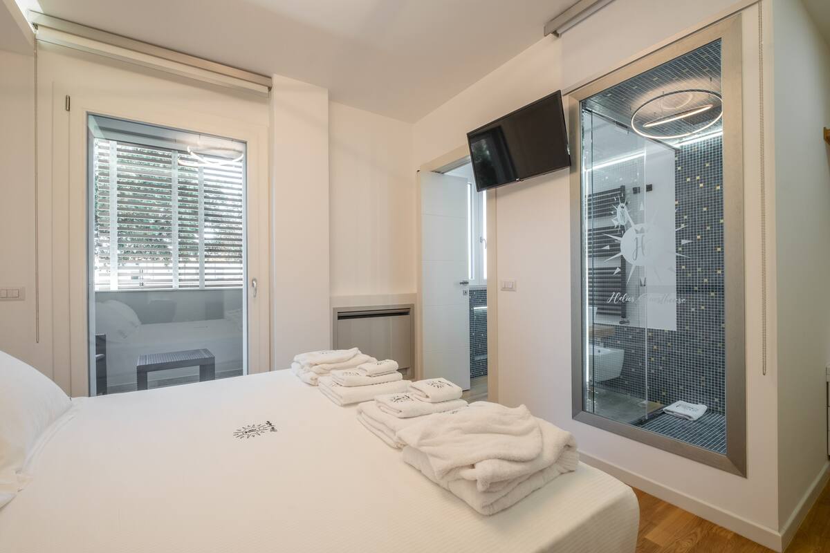 A hotel room with a bed, a glass shower enclosure, a TV mounted on the wall, and a view of the bathroom through the glass.
