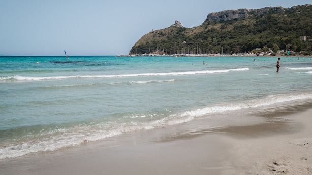 Une plage à l’eau turquoise et limpide, un rivage sablonneux et, au loin, une colline parsemée de végétation et de bâtiments.