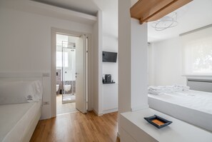 A modern bedroom with a wooden floor, a white bed, a TV on the wall, and a bathroom visible through an open door.