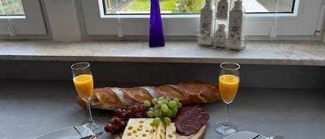 A breakfast setup with a croissant, cheese, grapes, and ham on a wooden board, accompanied by two glasses of orange juice, on a kitchen counter by a window.