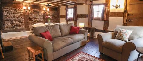 A cozy living room with exposed wooden beams, a stone wall, a fireplace, and a large red patterned rug.